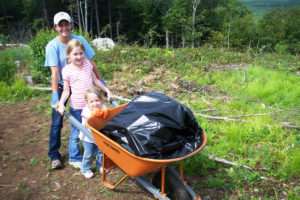 Photo of a young woman and two children holding onto a wheelbarrow. 
