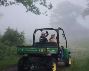 Photo of two Trail Crew members smiling and waving while driving a Gator, heading into the mist.