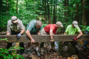 Photo of four men in the woods lifting a large piece of lumber.