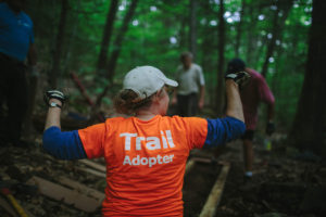 Photo of woman in the woods facing away from camera with a bright orange shirt that says "Trail Adopter."