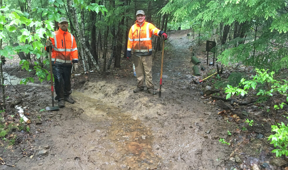 Two members of the Trail Crew wearing PPE standing next to a muddy trail with tools in hand.